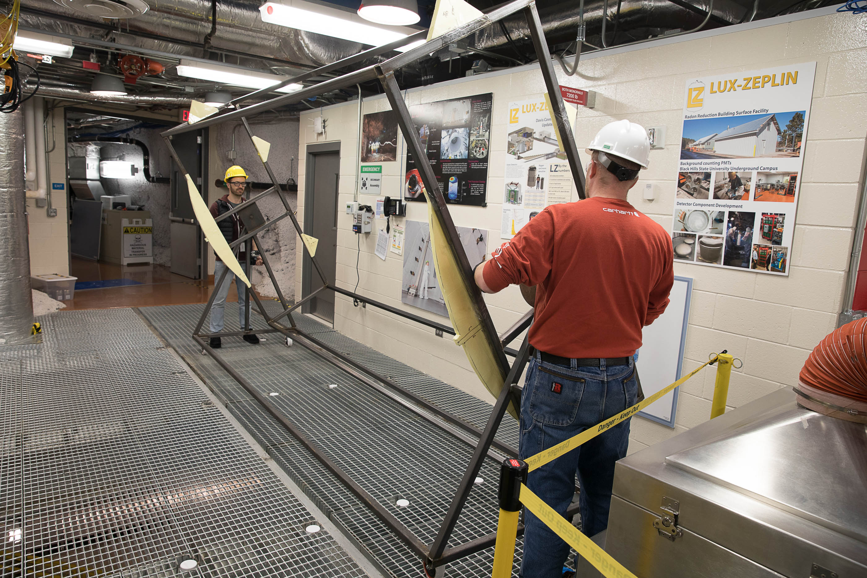 Two people stand at either end of a metal frame box with a rotary mechanism.