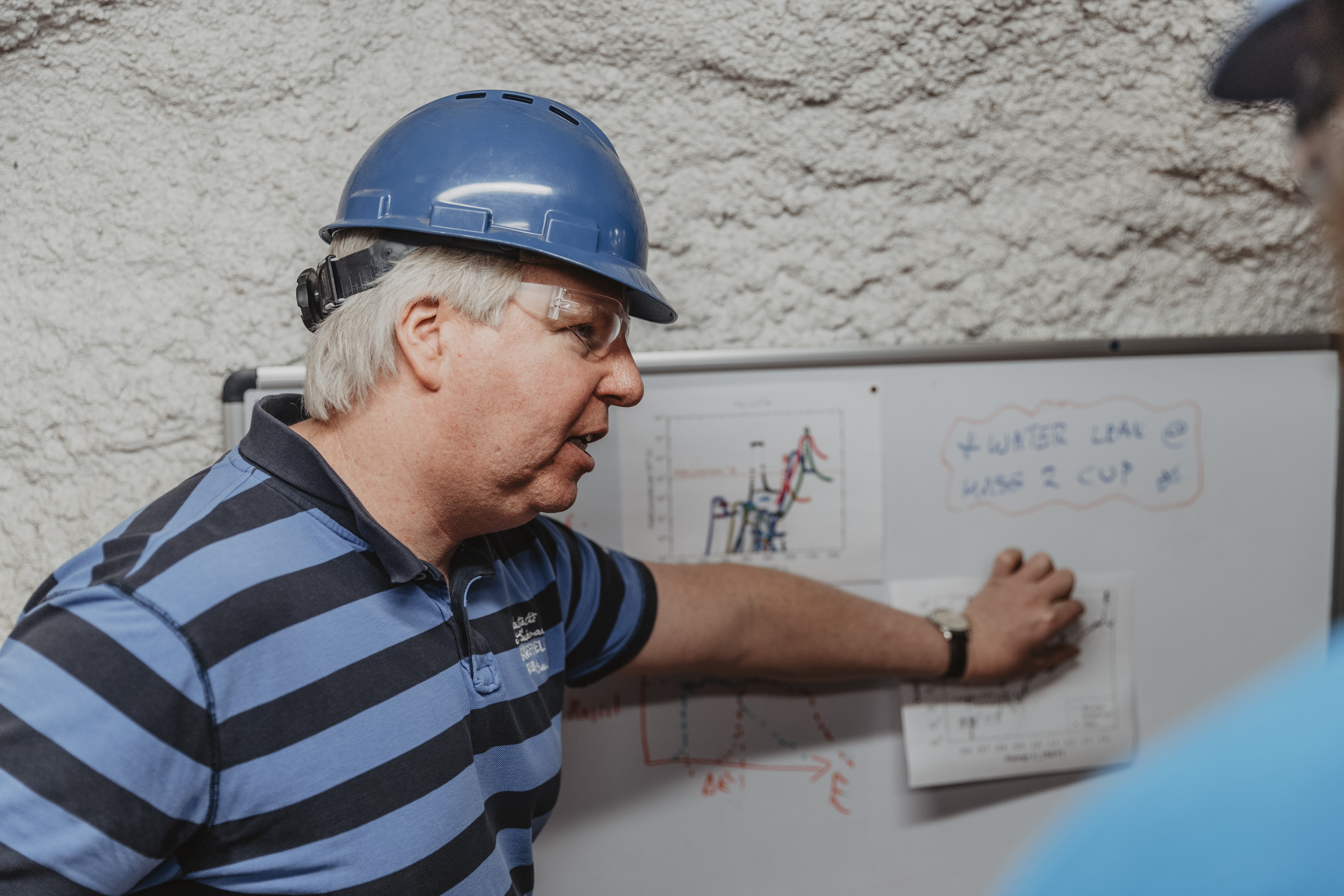 A researcher in a hard hat and safety glasses points to chart on the wall