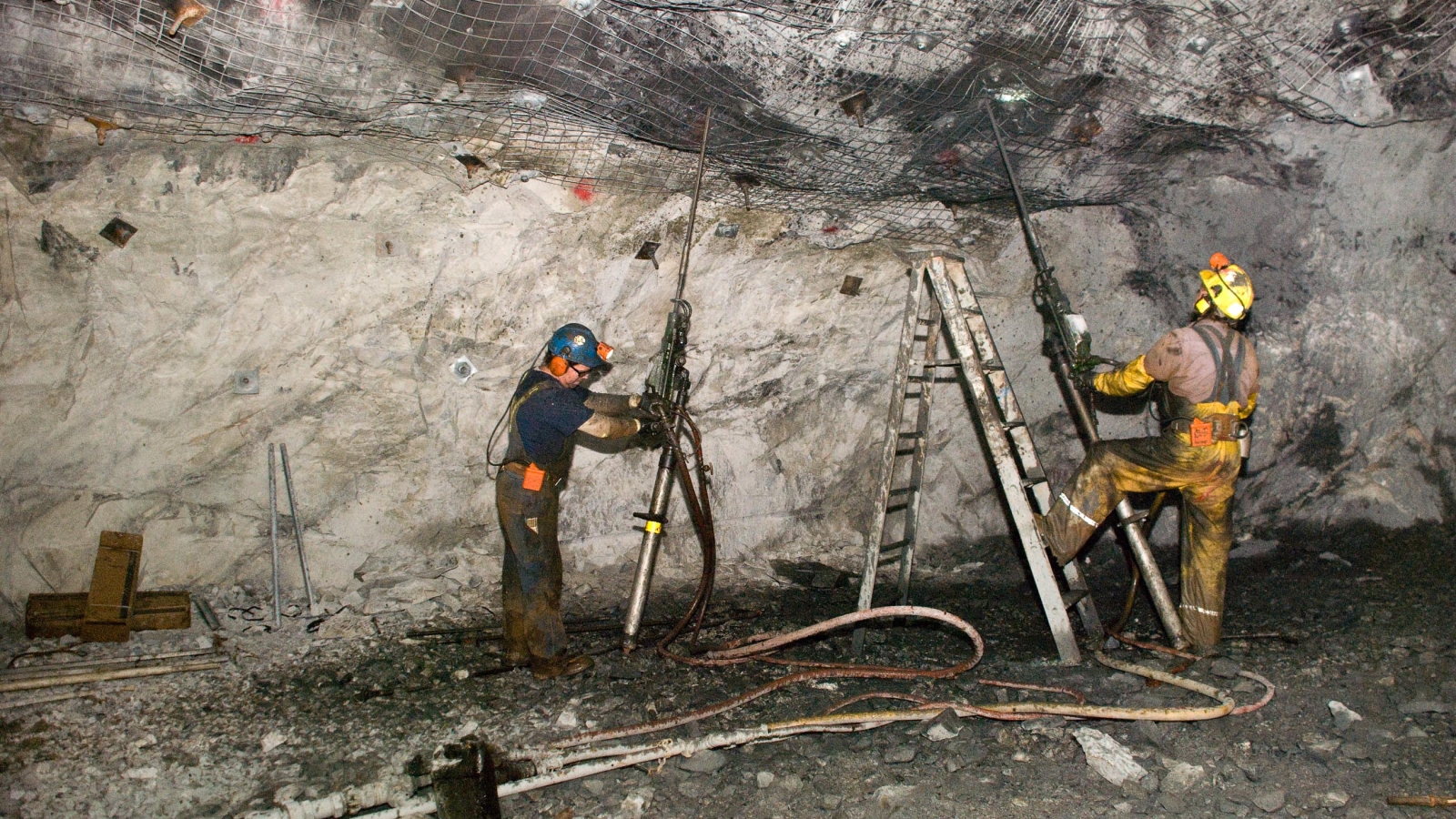 Two men use jackleg drills to install ground support in a cavern.
