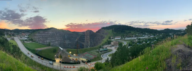 A crane lifts a large segment of steel conveyor truss in the air, over the highway. In the background, there is an open pit in the earth, created by strip mining.